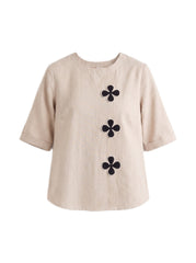 A beige short-sleeve top with three black four-petal flower motifs arranged vertically on the front, displayed against a solid white background.
