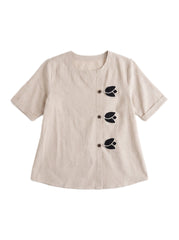 A beige short-sleeve linen top with three black embroidered leaf motifs and matching buttons down the front, displayed on a clean white background.