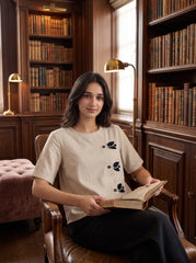 A woman sits in a leather armchair inside a warm, wood-paneled library, holding an open book. She is wearing a light beige linen short-sleeve top with black leaf embroidery and black trousers.