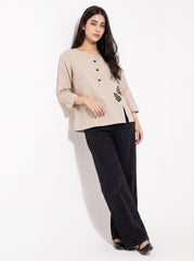 A woman modeling a beige linen three-quarter sleeve top with black buttons and a black leaf embroidery on the side, paired with black wide-leg trousers, against a plain white background.