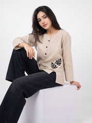 A woman sits on a white cube wearing a beige long-sleeve top with black buttons and a black leaf embroidery on the lower front, paired with dark jeans, against a plain white studio background.