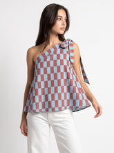 A woman modeling a one-shoulder peplum top with a dusty rose and light blue checkerboard pattern, paired with white pants, against a plain white studio background.