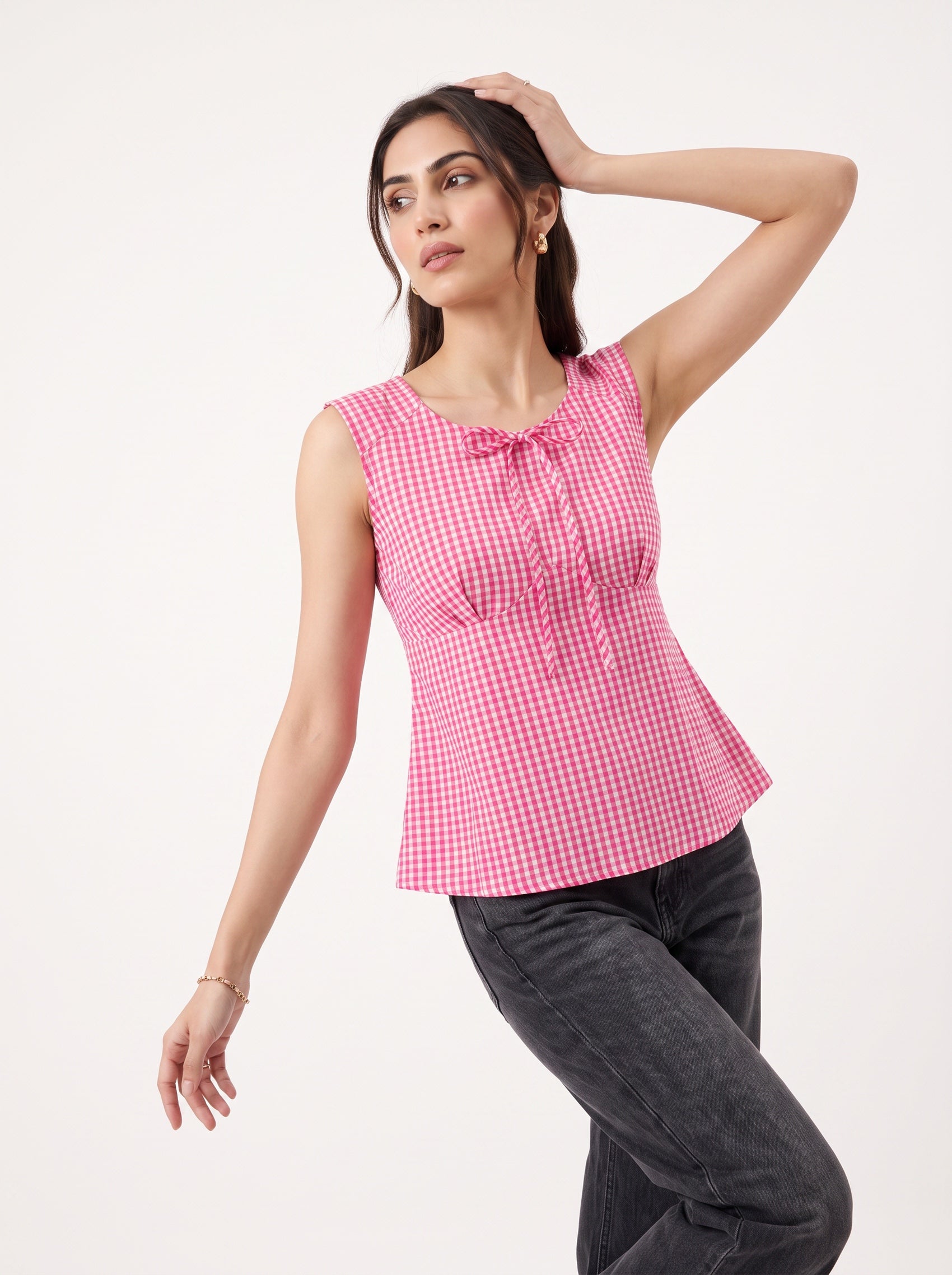 A woman modeling a sleeveless pink and white gingham top with a tie-neck detail, paired with dark gray jeans, against a plain white studio background.