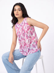 A woman wearing a sleeveless pink floral print top and light blue jeans, seated on a white stool against a plain white studio background.