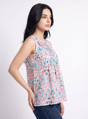 A woman wearing a sleeveless, pleated top with a pink, blue, and white geometric pattern, paired with dark blue jeans, photographed against a light gray studio background.