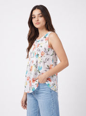 A woman wearing a sleeveless white top with a colorful floral print and light blue jeans, standing against a plain light gray studio background.