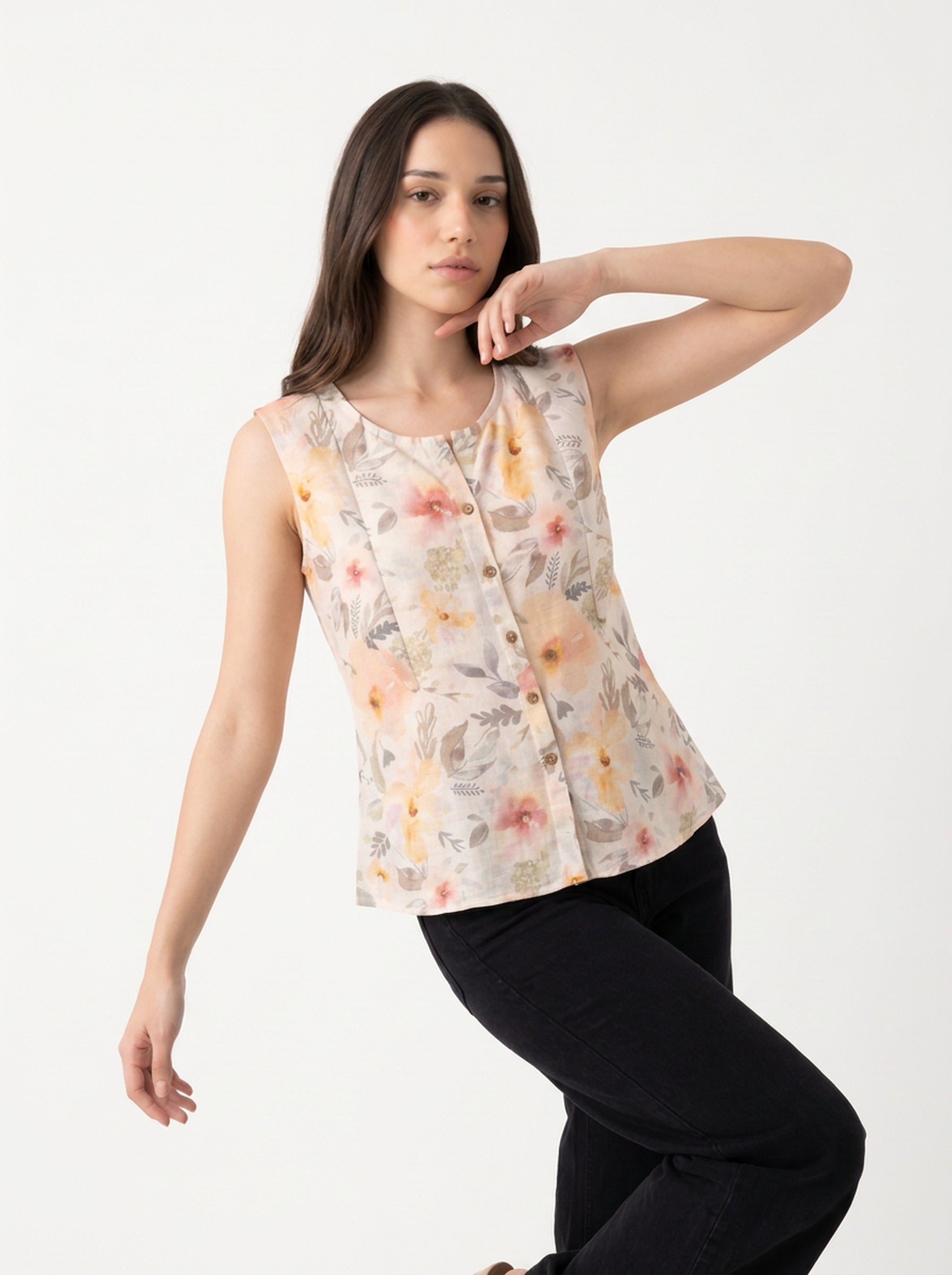 A woman modeling a sleeveless button-front top with a soft floral print in muted peach, yellow, and gray tones, paired with black pants, against a plain light background.
