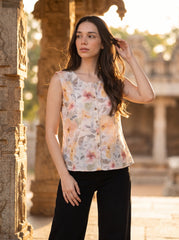 A woman wearing a sleeveless, button-front floral blouse with black pants, standing outdoors in front of a stone architectural structure during warm golden hour lighting.