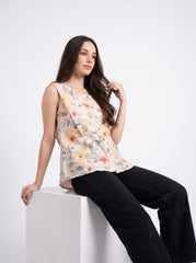 A woman with long dark hair sits on a white block, wearing a sleeveless beige blouse with a soft floral print in orange, pink, and green, paired with black wide-leg pants, against a plain white studio background.
