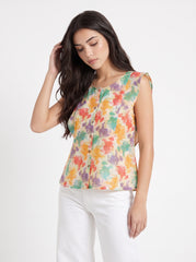 A woman wearing a sleeveless, button-front top with a colorful abstract floral print on a cream background, paired with white pants, photographed against a plain light gray studio background.