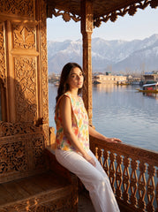 A woman wearing a sleeveless multicolor floral top and white pants sits on a carved wooden balcony overlooking a lake with mountains in the background.