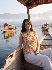 A woman wearing a sleeveless, button-front top with a colorful abstract floral print on a cream background, paired with white pants, photographed against a plain light gray studio background.