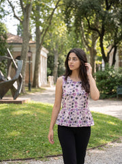 A woman wearing a sleeveless peplum top with a purple, pink, and black abstract floral pattern, paired with black pants, walking in a sunlit park with trees and a sculpture in the background.