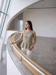 A woman stands on a curved indoor balcony wearing a sleeveless textured peplum top in a muted olive-beige tone with white pants, photographed in a modern minimalist interior with large windows and a concrete floor.