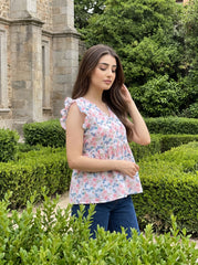 A woman wearing a sleeveless floral top with ruffle details and blue jeans, standing in a garden setting with green hedges and a stone building in the background.