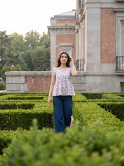 A woman stands in a formal garden wearing a light pink floral peplum top and dark blue wide-leg pants, with a historic brick building and trimmed hedges in the background.