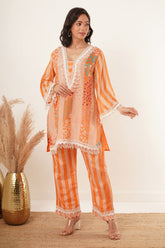 A woman is wearing a stylish orange mixed-print tunic set. The tunic features a combination of floral and striped patterns with fringe detailing along the neckline, hem, and sleeve edges. It is paired with matching striped trousers, creating a coordinated and vibrant ensemble. The outfit is complemented by neutral-toned heels, and the background includes decorative pampas grass in a golden vase, adding an elegant touch to the setting.