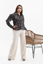 A woman stands in a studio wearing a black long-sleeve button-up shirt with a gray abstract leaf pattern, paired with cream wide-leg trousers and silver sandals. A woven rattan chair is positioned to her right against a plain white background.