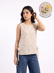 A woman modeling a sleeveless beige linen top with small embroidered flowers in yellow, pink, and green, paired with blue jeans. A circular inset in the top right highlights the floral embroidery detail.