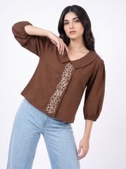 A woman modeling a brown cropped blouse with three-quarter puff sleeves, a wide collar, and white floral embroidery down the front, paired with light blue jeans against a plain white background.