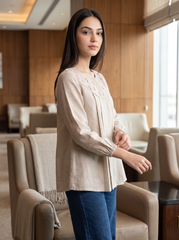 A woman wearing a beige long-sleeve linen blouse with pink and green floral embroidery on the chest, paired with blue jeans, standing in a modern lounge setting with beige armchairs.