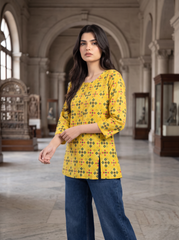 A woman modeling a yellow three-quarter sleeve kurta with a green and orange geometric pattern, paired with dark blue jeans, standing in a grand, arched hall resembling a museum or library.