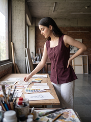 A woman in a studio setting wearing a sleeveless maroon top and white pants, standing at a wooden table covered with papers, swatches, and art supplies.