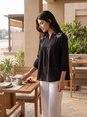 A woman stands at an outdoor patio table wearing a black long-sleeve top with delicate white floral embroidery on the chest and white trousers. She is reaching for a plate of fruit on a wooden table set with teacups and saucers.