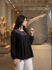 A woman wearing a black long-sleeve blouse with intricate silver embroidery around the neckline, paired with white pants, standing in a museum setting next to a glass display case containing an ancient stone tablet.