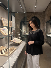 A woman wearing a black long-sleeve blouse with a detailed silver-gray embroidered yoke and white trousers stands in a museum gallery next to a glass display case containing historical books and chess pieces.