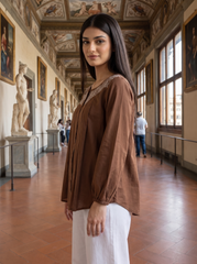 A woman wearing a brown long-sleeve blouse with lace trim and white pants stands in a grand museum hallway with classical statues and paintings.
