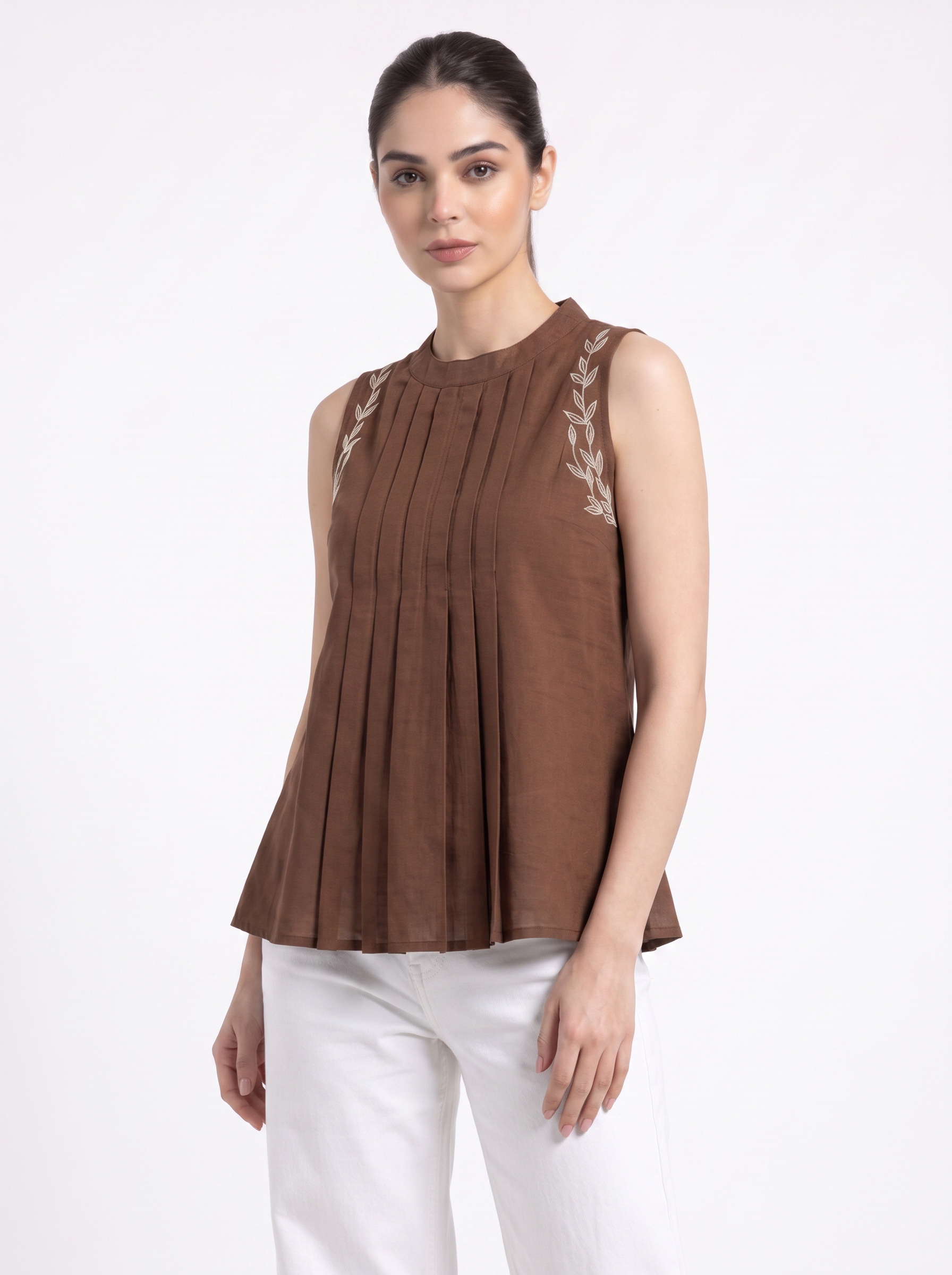 A woman modeling a sleeveless brown pleated top with white leaf embroidery on the shoulders, paired with white pants, against a plain white studio background.