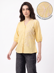 A woman wearing a light yellow linen blouse with white hand-embroidered floral patterns on the front, paired with dark gray jeans. A circular inset in the top right corner highlights the detailed hand embroidery with the text 'HAND EMBROIDERY'.