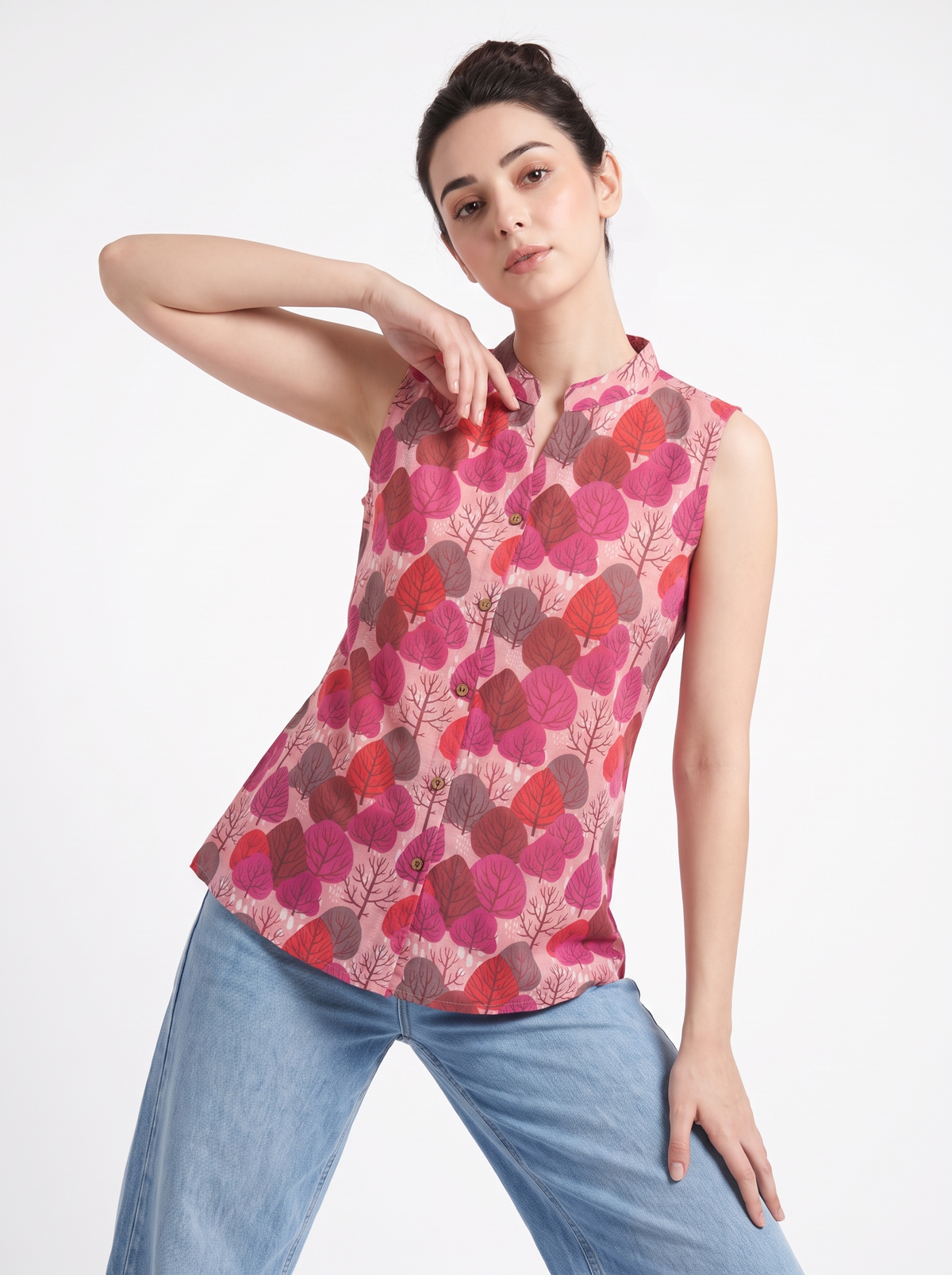 A woman modeling a sleeveless, mandarin-collar blouse with a pink, red, and brown leaf pattern, paired with light blue jeans, against a plain white studio background.
