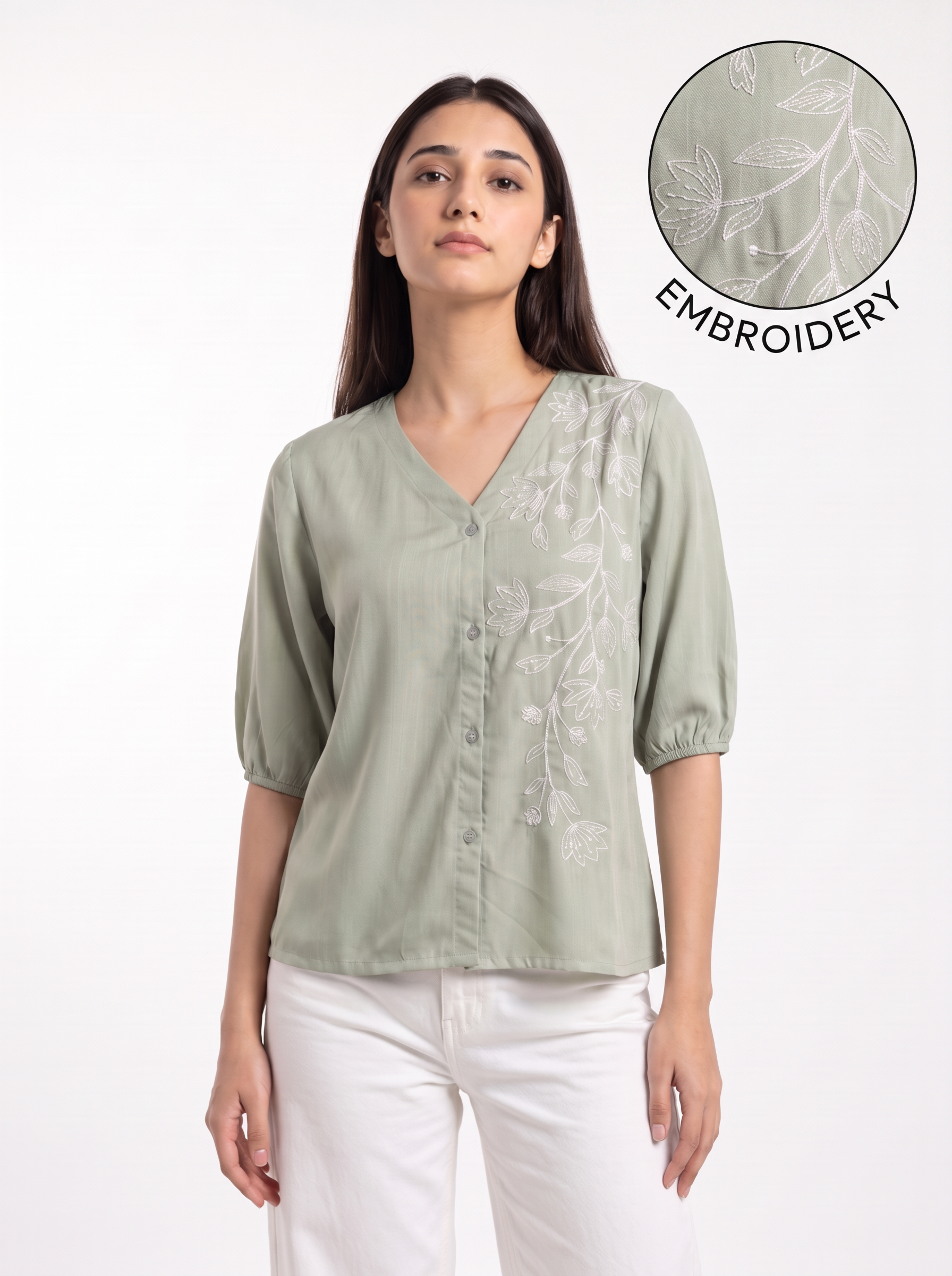 A woman wearing a sage green short-sleeve blouse with white floral embroidery on the front, paired with white pants. A circular inset in the top right shows a close-up of the embroidery detail with the text 'EMBROIDERY'.