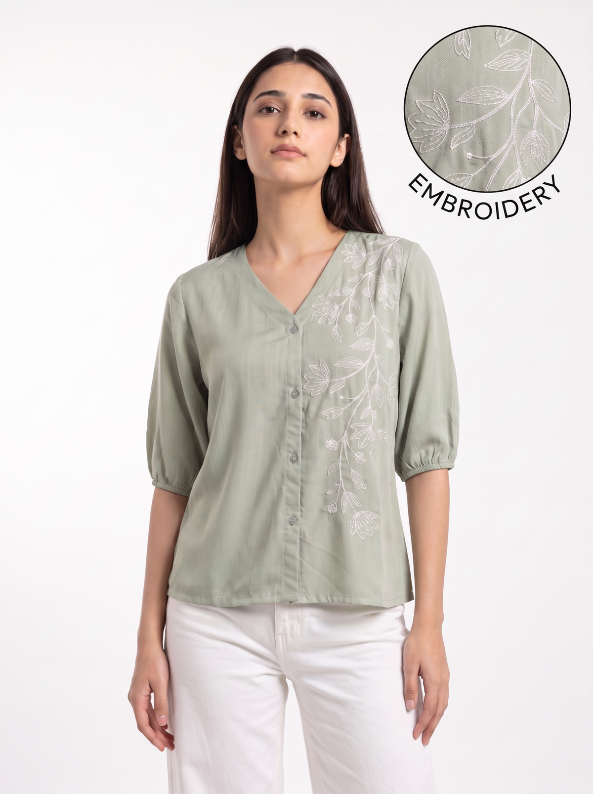 A woman wearing a sage green short-sleeve blouse with white floral embroidery on the front, paired with white pants. A circular inset in the top right shows a close-up of the embroidery detail with the text 'EMBROIDERY'.