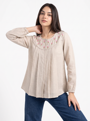 A woman modeling a light beige linen long-sleeve blouse with pink floral embroidery around the neckline, paired with blue jeans, against a plain white background.