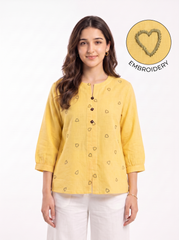 A woman modeling a yellow three-quarter sleeve top with small embroidered heart motifs, paired with white pants, against a plain white background. An inset circle in the top right shows a close-up of the yellow fabric with a stitched heart and the word 'EMBROIDERY'.