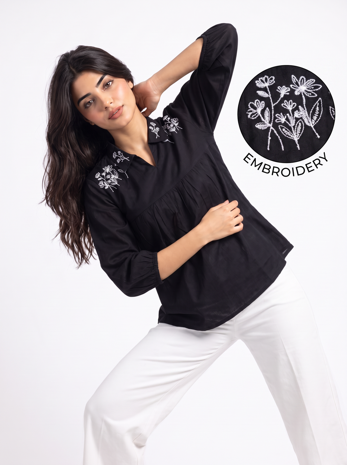 A woman walking on a sandy beach wearing a black long-sleeve blouse with white floral embroidery on the shoulders and white pants, with the blue sea and resort buildings in the background.