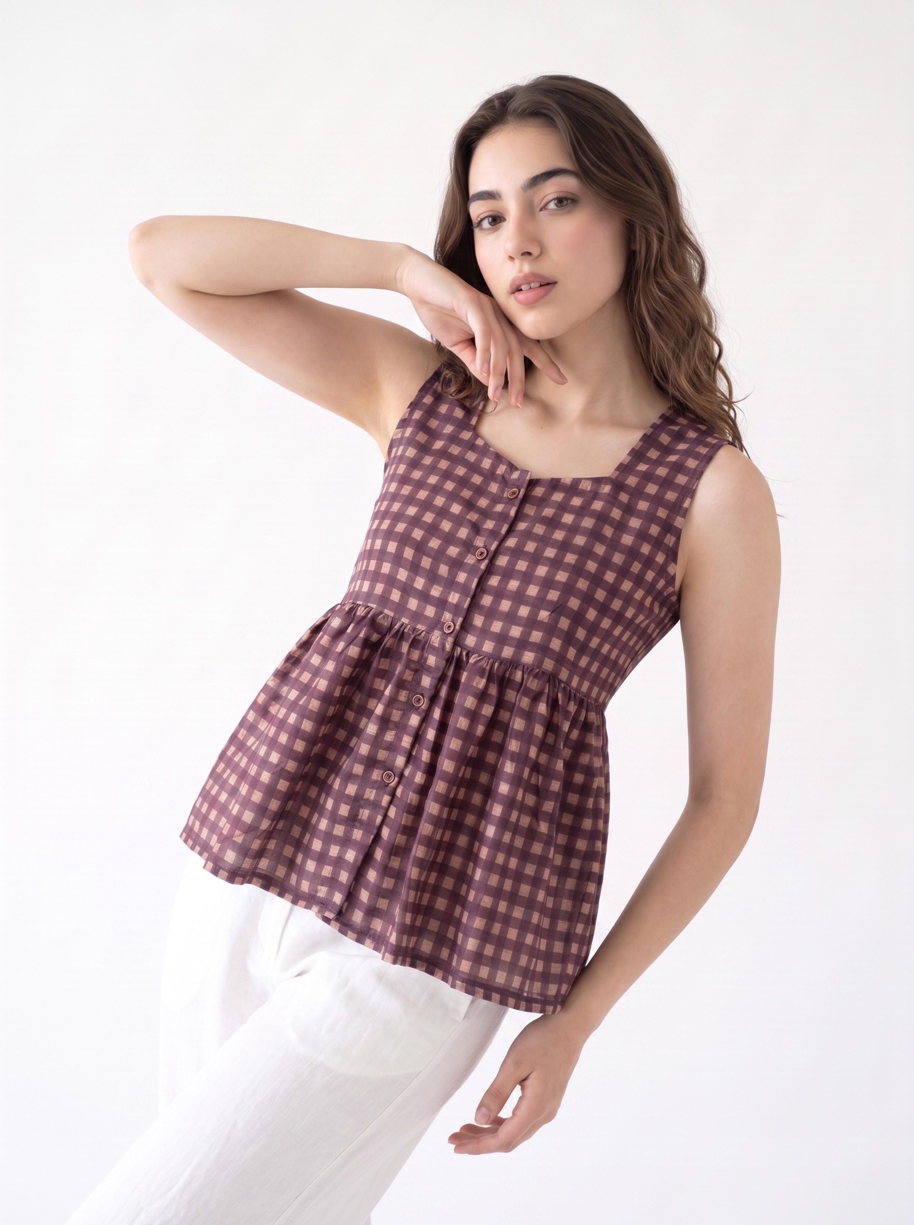 A woman modeling a sleeveless peplum top with a brown and pink gingham pattern, paired with white pants, against a plain white background.