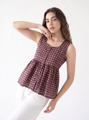 A woman modeling a sleeveless peplum top with a brown and pink gingham pattern, paired with white pants, against a plain white background.