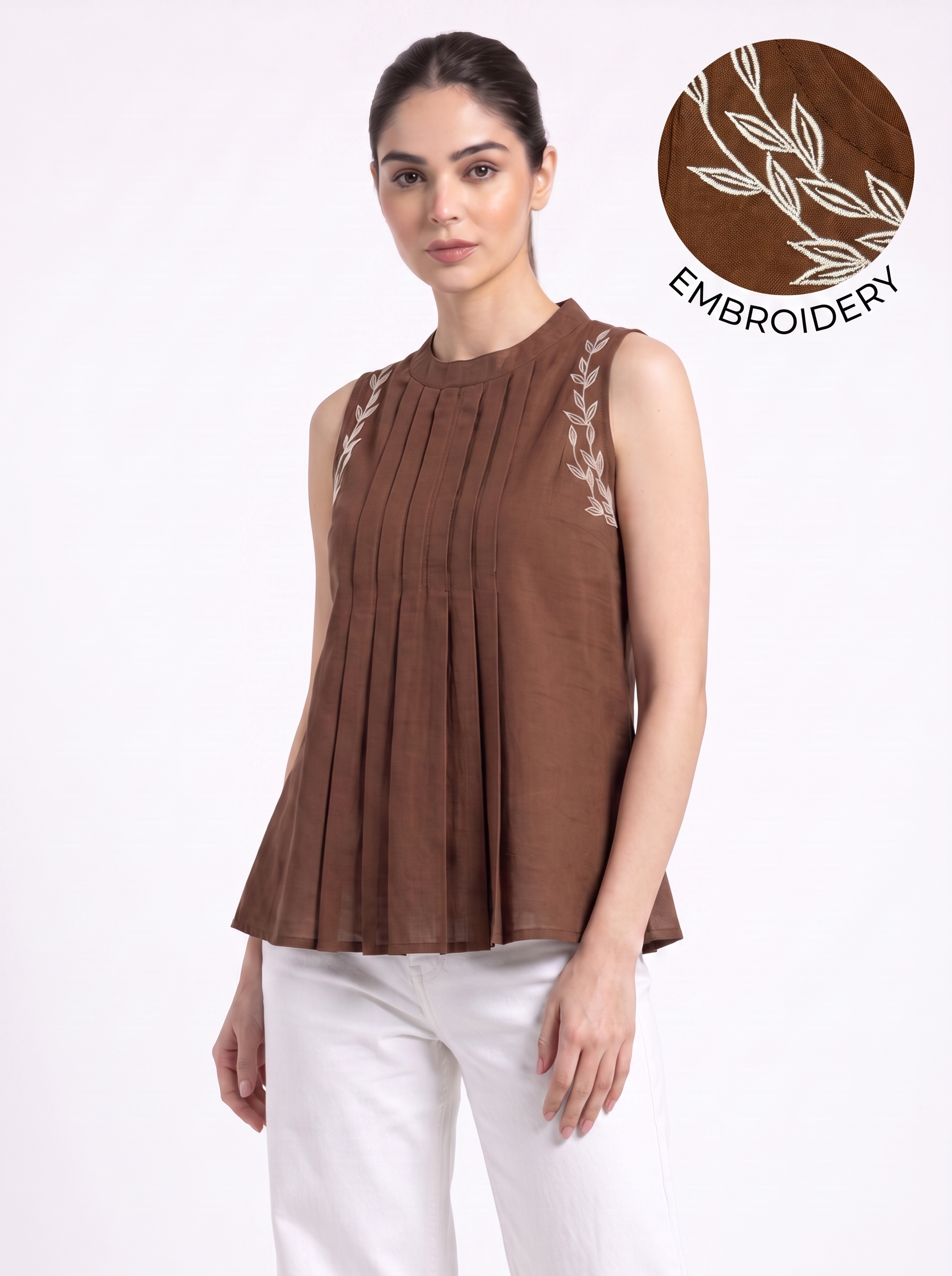 A woman modeling a sleeveless brown pleated top with white embroidered leaf details on the shoulders, paired with white pants, against a plain white background. An overlay graphic in the top right shows a close-up of the brown fabric with white embroidery and the word 'EMBROIDERY'.