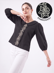 A woman modeling a black long-sleeve blouse with white floral embroidery down the front, paired with white pants. A circular inset in the top right shows a close-up of the white floral embroidery on black fabric with the word 'EMBROIDERY'.