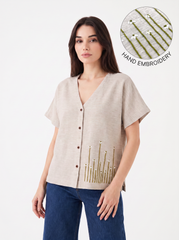 A woman modeling a light beige linen short-sleeve button-up top with hand-embroidered green and white floral details near the hem, paired with blue jeans. An inset circle in the top right highlights the hand embroidery.