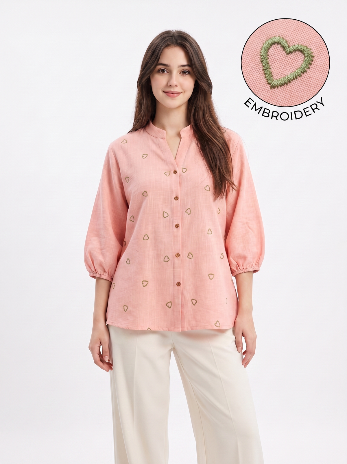 A woman modeling a light pink linen button-up blouse with small embroidered heart motifs, paired with cream-colored pants, against a plain white studio background. An inset circle in the top right highlights the green and pink embroidery detail with the text 'EMBROIDERY'.