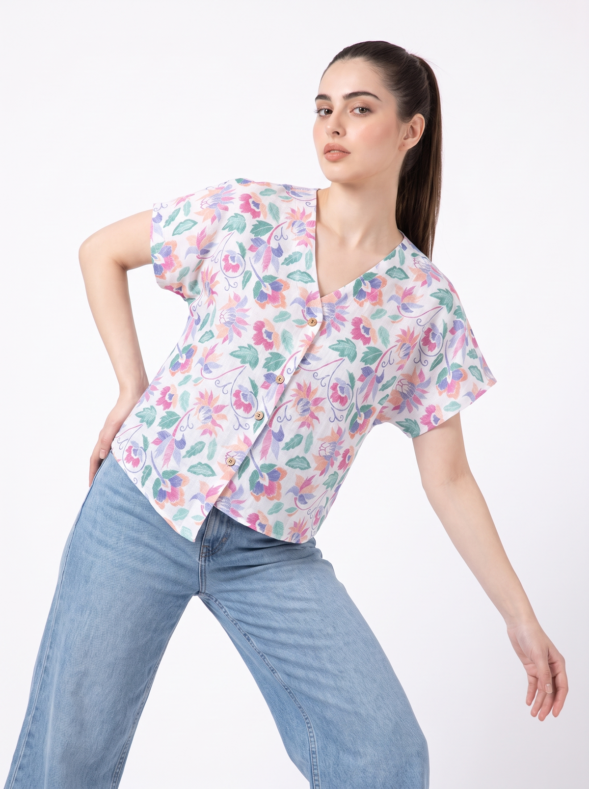 A woman modeling a short-sleeve V-neck button-up scrub top with a colorful floral print, paired with light blue jeans, against a plain white background.