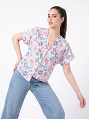 A woman modeling a short-sleeve V-neck button-up scrub top with a colorful floral print, paired with light blue jeans, against a plain white background.