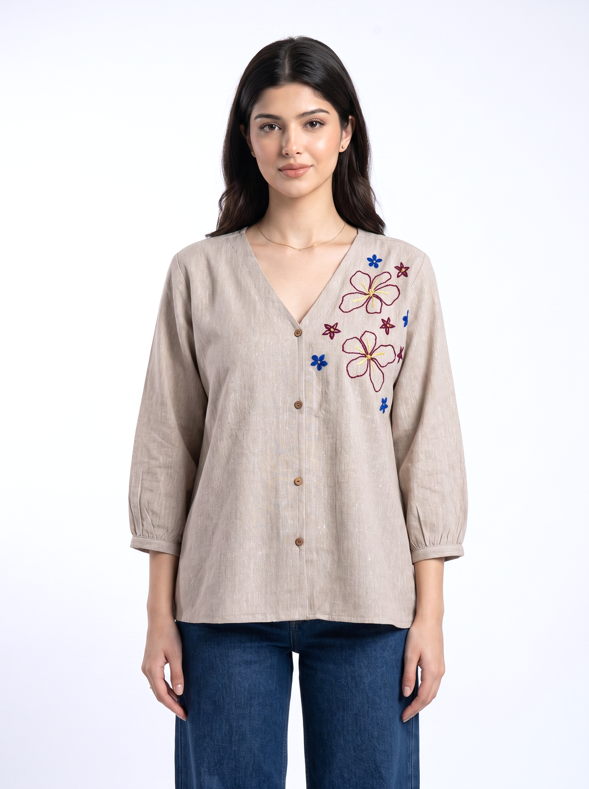 A woman modeling a beige linen button-up blouse with colorful floral embroidery on the chest, paired with blue jeans, against a plain white studio background.