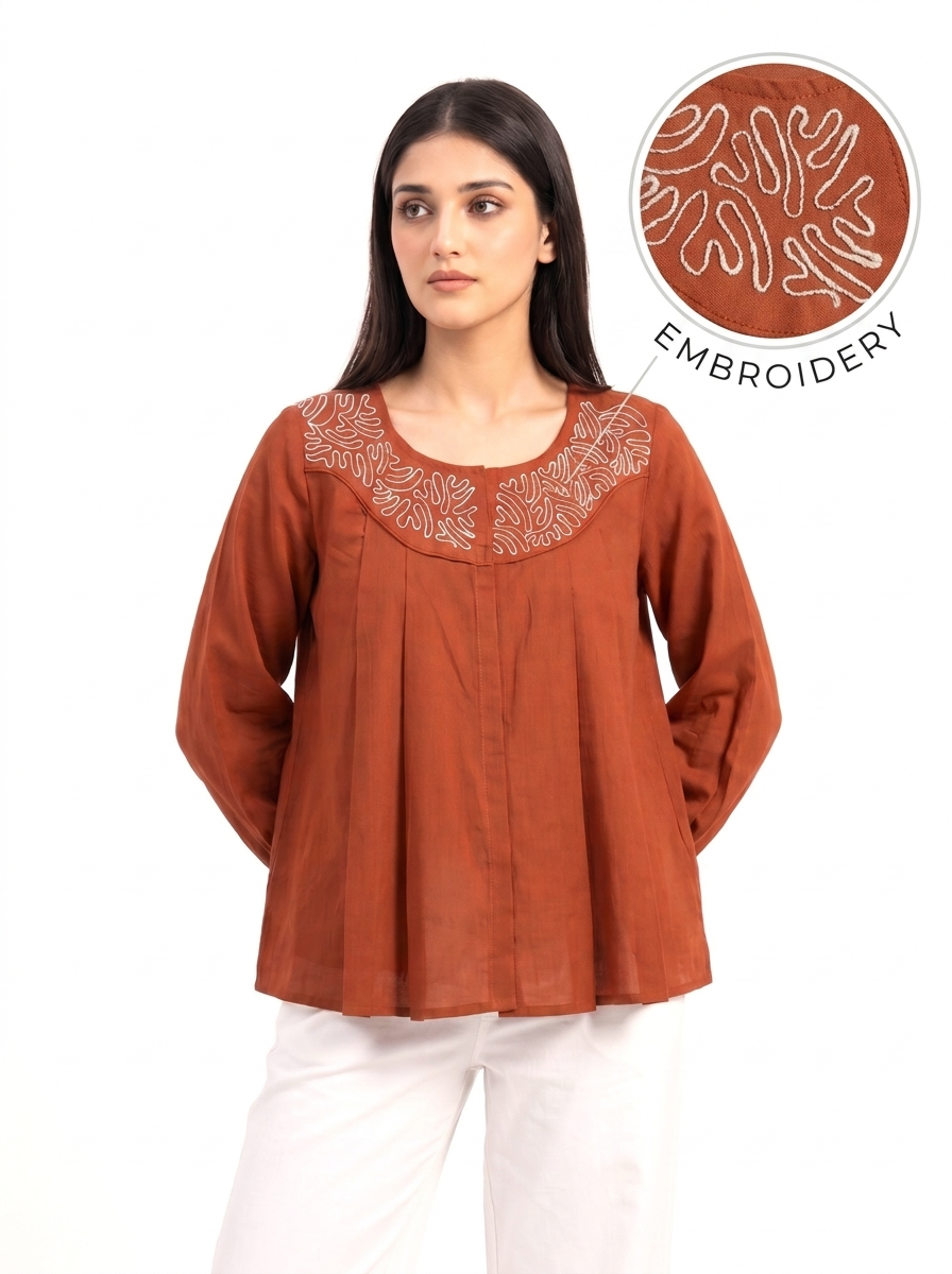 A woman wearing a rust-orange long-sleeve top with white embroidered detailing around the neckline, paired with white pants. An inset circle in the upper right highlights the embroidery texture with the word 'EMBROIDERY' on a white background.