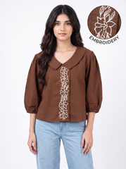 A woman wearing a brown long-sleeve blouse with white floral embroidery down the front, paired with light blue jeans. A circular inset in the top right shows a close-up of the white embroidery detail.